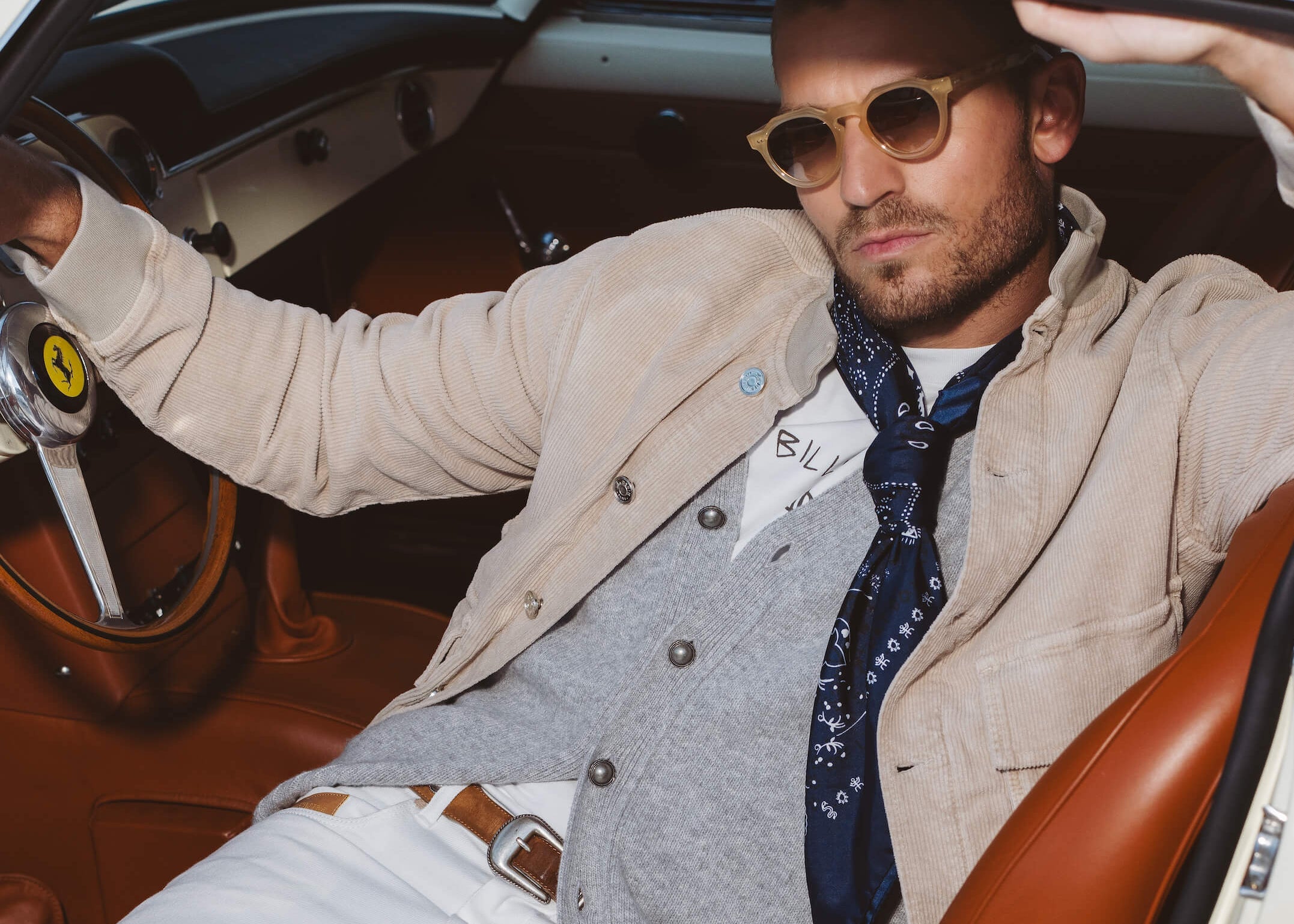 Mannequin portant cardigan gris, jeans blanc, veste en velours et ceinture marron assis dans une ferrari classique