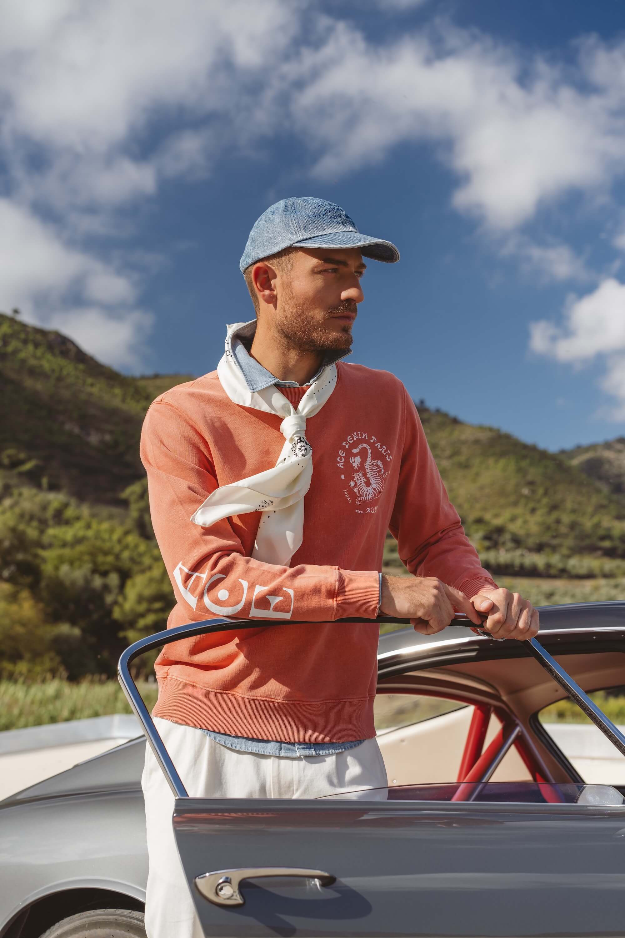 Mannequin portant un pull orange avec foulard blanc et pantalon chino blanc devant une Ferrari 250