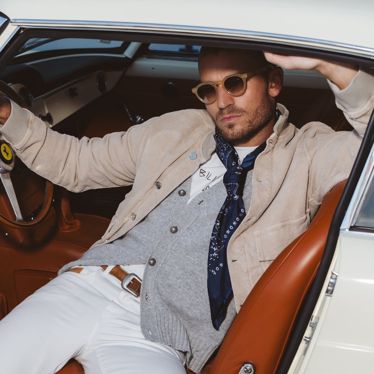 Mannequin portant un cardigan gris, tee-shirt billionaire, jeans blanc, veste en velours beige et ceinture marron assis dans une ferrari classique